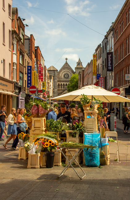 A lively street scene on a sunny day featuring a flower stall with fresh floral arrangements displayed in wooden crates and vases. The display includes vibrant sunflowers, pink roses, lilies, and vari