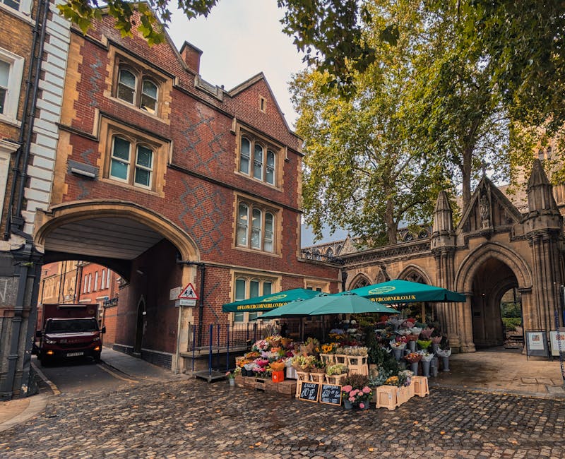 A street scene in St Johns Wood, featuring a blossom-filled flower stall set up under a green parasol on a cobbled pavement near a historic red brick building with an arched gateway and Gothic-style s