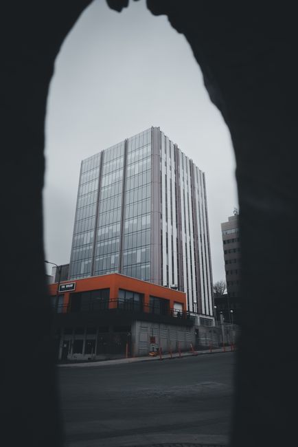 The image shows a bouquet of fresh flowers held by a person with dark clothing, positioned in front of a modern high-rise building with glass panels, set against a gray, overcast sky. The bouquet feat