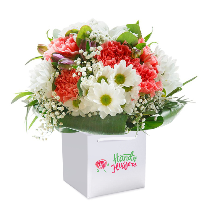 Positioned upright inside a gleaming square white gift bag, this exuberant bouquet reflects both the vibrance and tranquillity characteristic of St John's Wood. The focal point is a rich blend of coral-pink and peach carnations; their plush, ruffled petals wash the arrangement in warmth, much like the sunset filtering through the mature trees on Acacia Road. Interspersed between these textured blooms, bright white daisy-chrysanthemums stand proud, their juicy lime-green centers offering a splash of fresh energy. Sprays of delicate white gypsophila weave their way among the flowers, creating a diaphanous veil that softens the entire display. Scattered among the blossoms, closed buds tinged with purple and blush pink add depth and a hint of anticipation, echoing the layered charm of St John's Wood's hidden avenues. Wrapping the ensemble, glossy, deep emerald foliage arcs wide to form a natural, supportive frame, grounding the arrangement with robust life. The overall effect is full-bodied and generous, designed to evoke joy and elegance in equal measure. The practical ribbon handles on the white bag provide ease for same-day gifting or doorstep delivery anywhere from Avenue Road to Maida Vale. Bathed in gentle light against a clear white field, this florist-made bouquet becomes a beacon of quality, artistry, and the personal touch that is the hallmark of St John's Wood's local flower scene.
